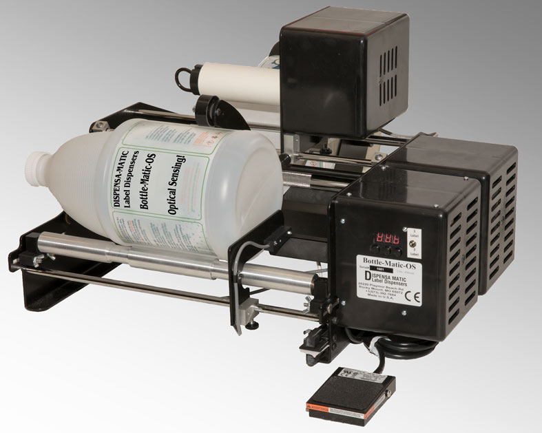 Bottle Matic OS label dispenser with a large white plastic bottle mounted on a compact black metal frame. The device features electronic controls and a digital display, designed for dispensing and applying labels onto bottles or containers in manufacturing or packaging environments. Its efficient, automated operation helps streamline labeling processes for bottles in industrial settings.